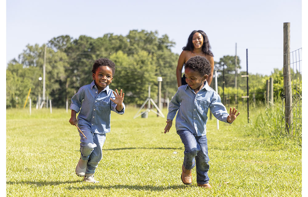 Twin boys playing with their mom, running in a grassy field with trees behind them.